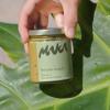 a handing coming out of the top of the frame holding a hawaiian macadamia nut butter jar in a pistachio flavor with a gold lid and the maka logo and packaging design, sitting against a background of a large green palm leaf
