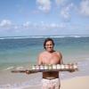 an excited surfer on a sunny day in a tropical beach holding a wooden palette with a line of maka products in various colors