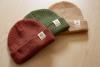 Three beanies laid out on a wooden table with tags that have a pinecone logo on it. One beanie is red, one is green and one is beige.