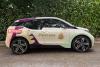 Right side view of a BMW i3 parked in front of a green hedge wrapped in graphics for Pinecone Childcare in Portland, Oregon. It is mostly beige with minty green, pink and dark purple shapes. It has a pinecone logo on it with the slogan "Childcare done nicely" and a red shape with a qr code and text that reads "Register now!" and "pinecone-childcare.com".