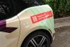 Right back side view of a BMW i3 in front of a green hedge wrapped in graphics for Pinecone Childcare in Portland, Oregon. It is mostly beige with dark purple and minty green shapes. It has a red shape with a qr code and text that reads "Register now!" and "pinecone-childcare.com".