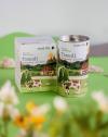 Packaging design for "Pure Tea" with the flavor "Traudl" labeled as "Almkräuter - Alpine Herbs." The packaging includes a box and a cylindrical tin. Both feature an illustrated scenic landscape with an Alpine theme, showcasing green hills, trees, a traditional chalet-style building, mountains, and a cow in the foreground. The background has soft, earthy tones and a light green surface, adding to the natural and fresh vibe of the packaging.